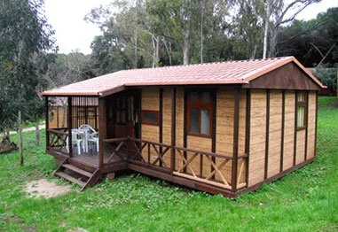 Small rustic wooden cabin with a red tile roof, front porch, railing, and surrounding green grass and trees