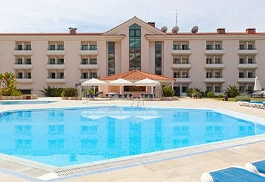Large outdoor swimming pool in front of a four-story resort hotel with umbrellas and lounge chairs
