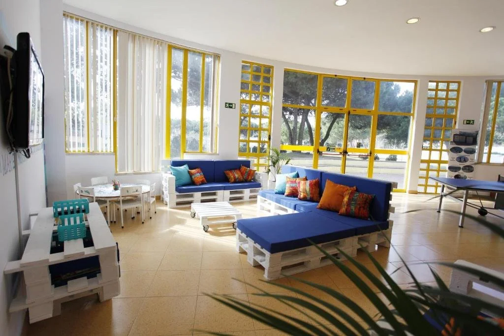 Bright modern lounge with blue pallet sofas, colorful cushions, large yellow-framed windows, and a pool table