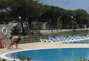 Outdoor swimming pool with blue lounge chairs, fenced tennis courts, and a person crouching on the pool deck
