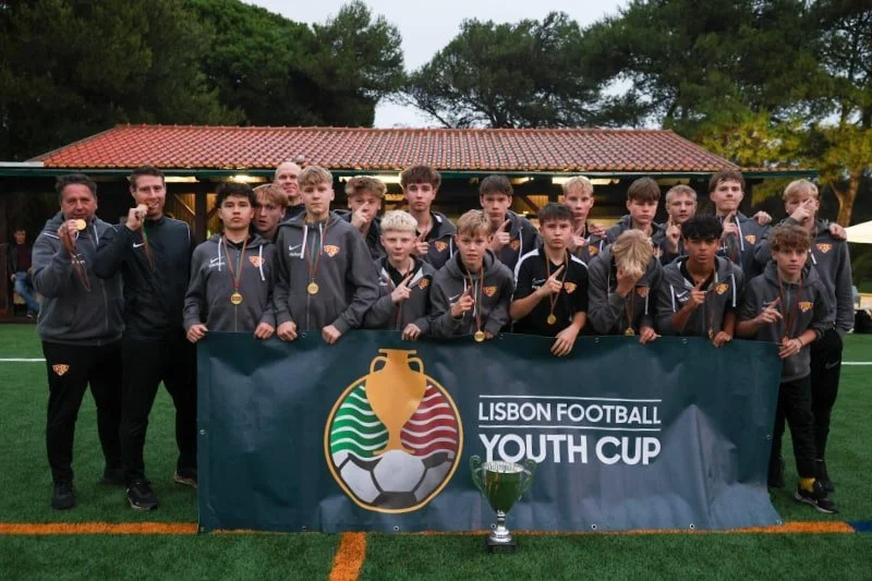 Youth soccer team posing with medals behind a Lisbon Football Youth Cup banner and trophy