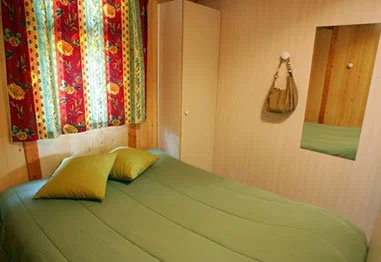Cozy bedroom with a green bedspread, yellow pillows, colorful floral curtains, wardrobe, handbag, and wall mirror