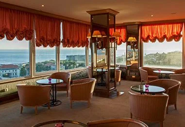 Elegant hotel lounge with red drapes, wicker chairs, tables, and panoramic ocean and coastal city views