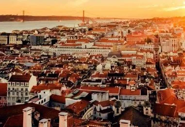 Aerial view of Lisbon’s red-roofed historic cityscape at sunset, with the Tagus River and 25 de Abril Bridge in the background.