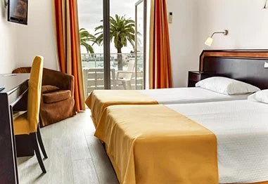 Hotel room with two beds, orange curtains, seating area, and balcony overlooking palm trees