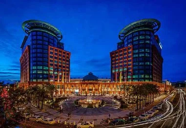 Nighttime aerial view of twin modern office towers surrounding a brightly lit circular plaza and curving roads