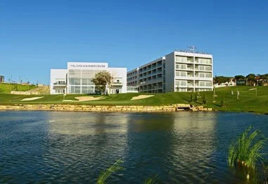 Modern waterfront hotel and conference center beside a landscaped lawn and lake under a clear blue sky