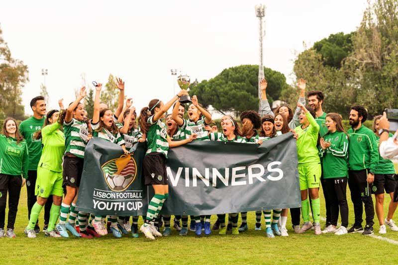 Youth soccer team celebrating a tournament win with a trophy and “Winners” banner at the Lisbon Football Youth Cup