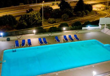 Illuminated outdoor swimming pool with blue lounge chairs beside a curving highway at night