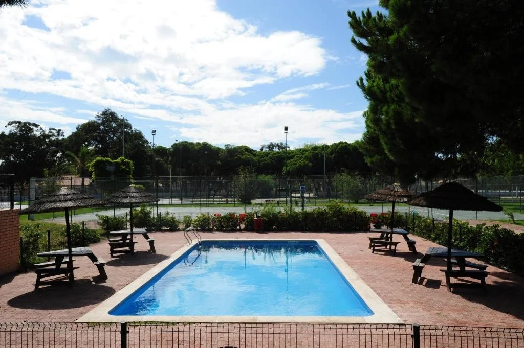 Outdoor swimming pool with thatched umbrellas, picnic tables, and fenced tennis courts under a partly cloudy sky