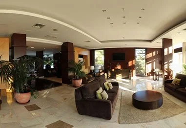 Modern hotel lobby with sofas, a round coffee table, potted plants, marble floors, and large sunlit windows
