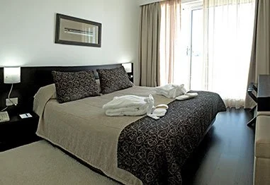 Hotel bedroom with a queen bed, patterned pillows and bedspread, folded towels, nightstand, and bright balcony doors.