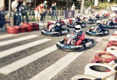 Go-kart racers speeding across a track near a striped crosswalk, with tire barriers and spectators in the background