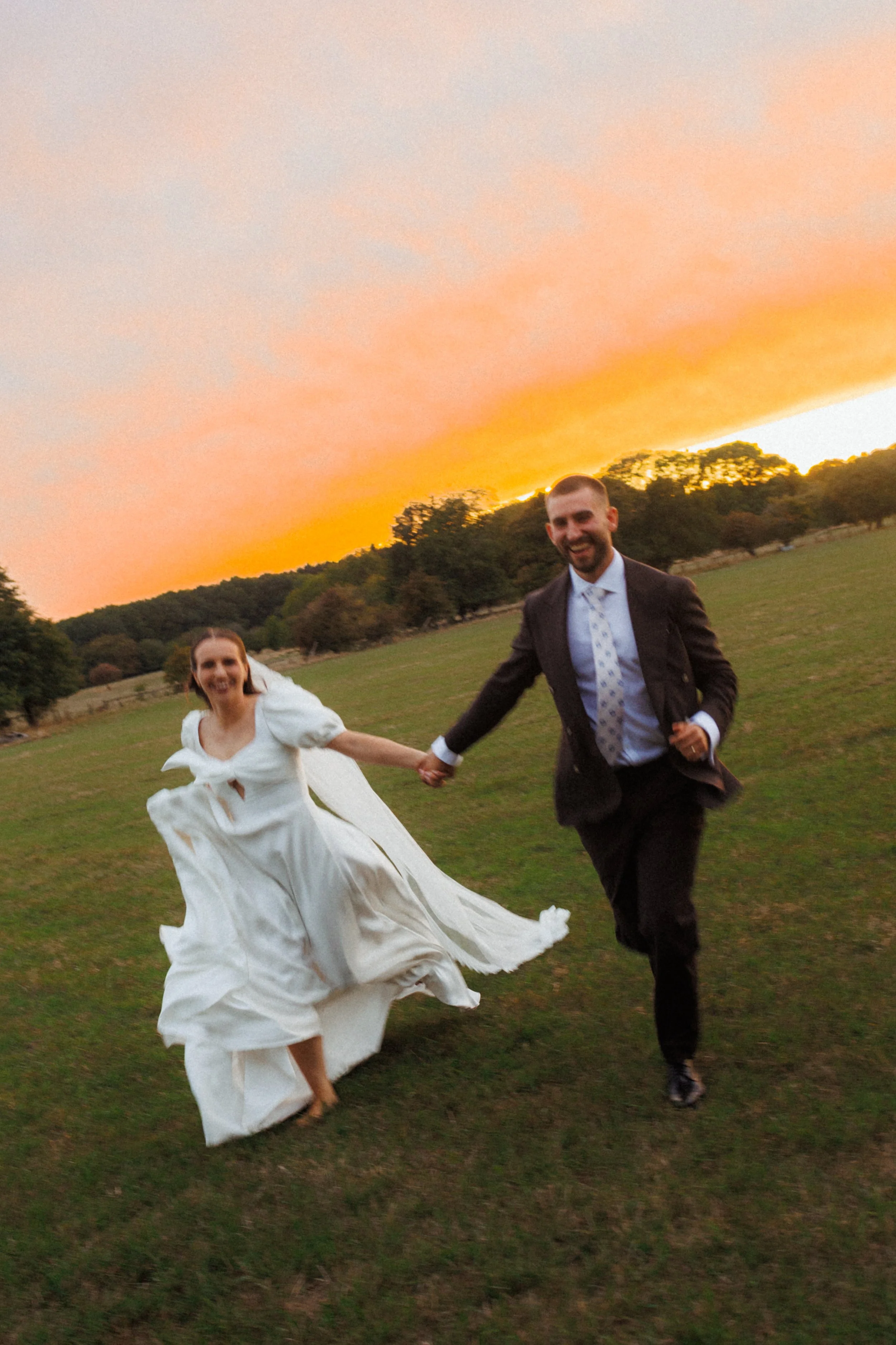 A bride and groom holding hands and running across a grassy field during sunset, with trees in the background.