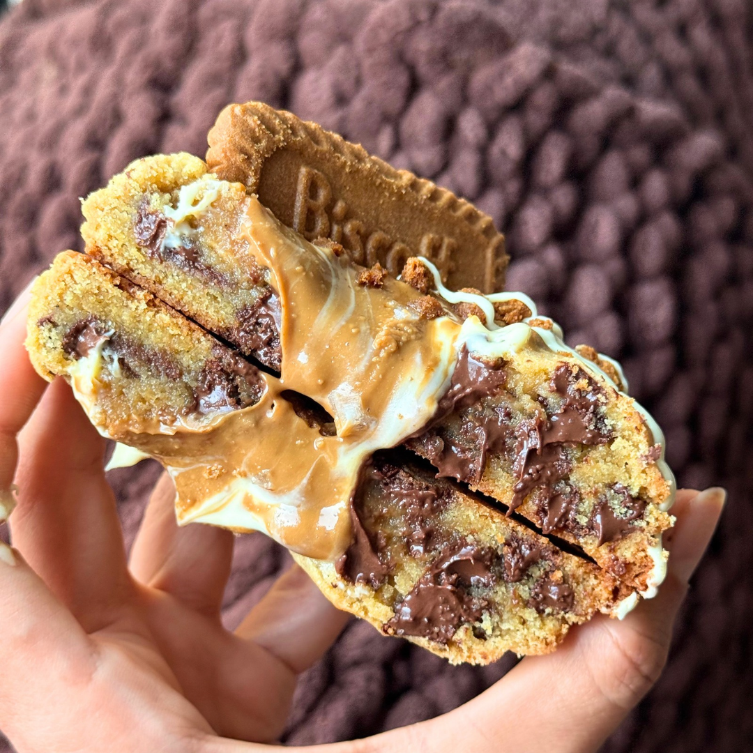 Close-up of a cookie sandwich with chocolate chips, caramel, and white chocolate drizzle, held in hand against a brown textured blanket background.