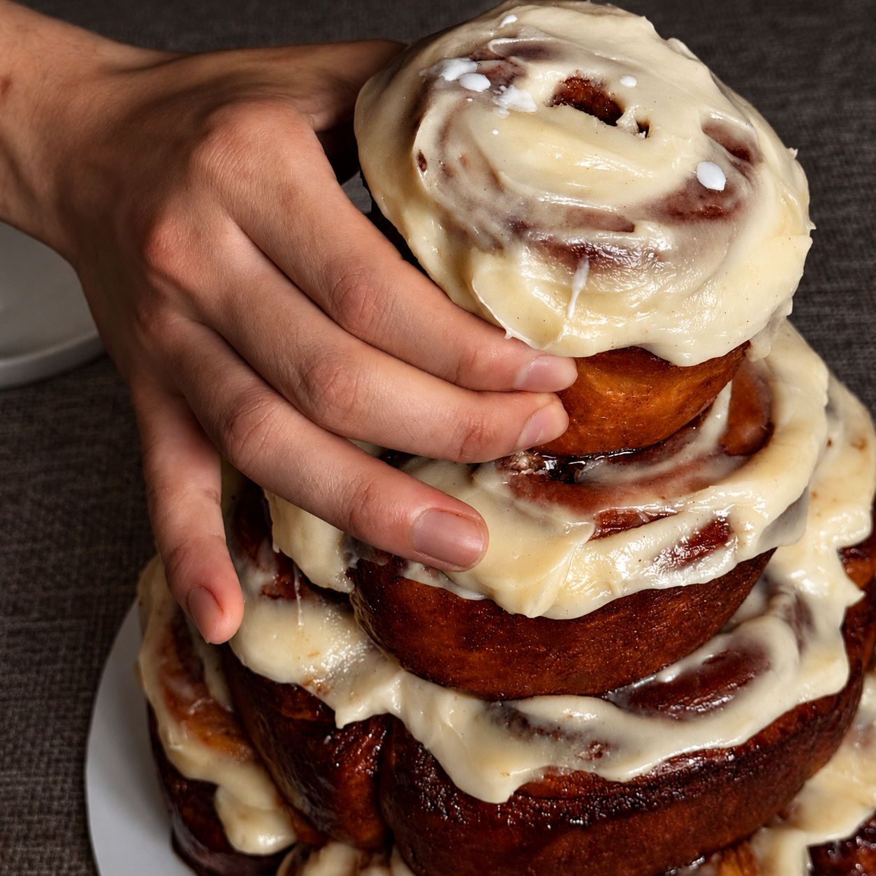 Close-up of a person's hand touching a cinnamon roll cake topped with cream cheese frosting.