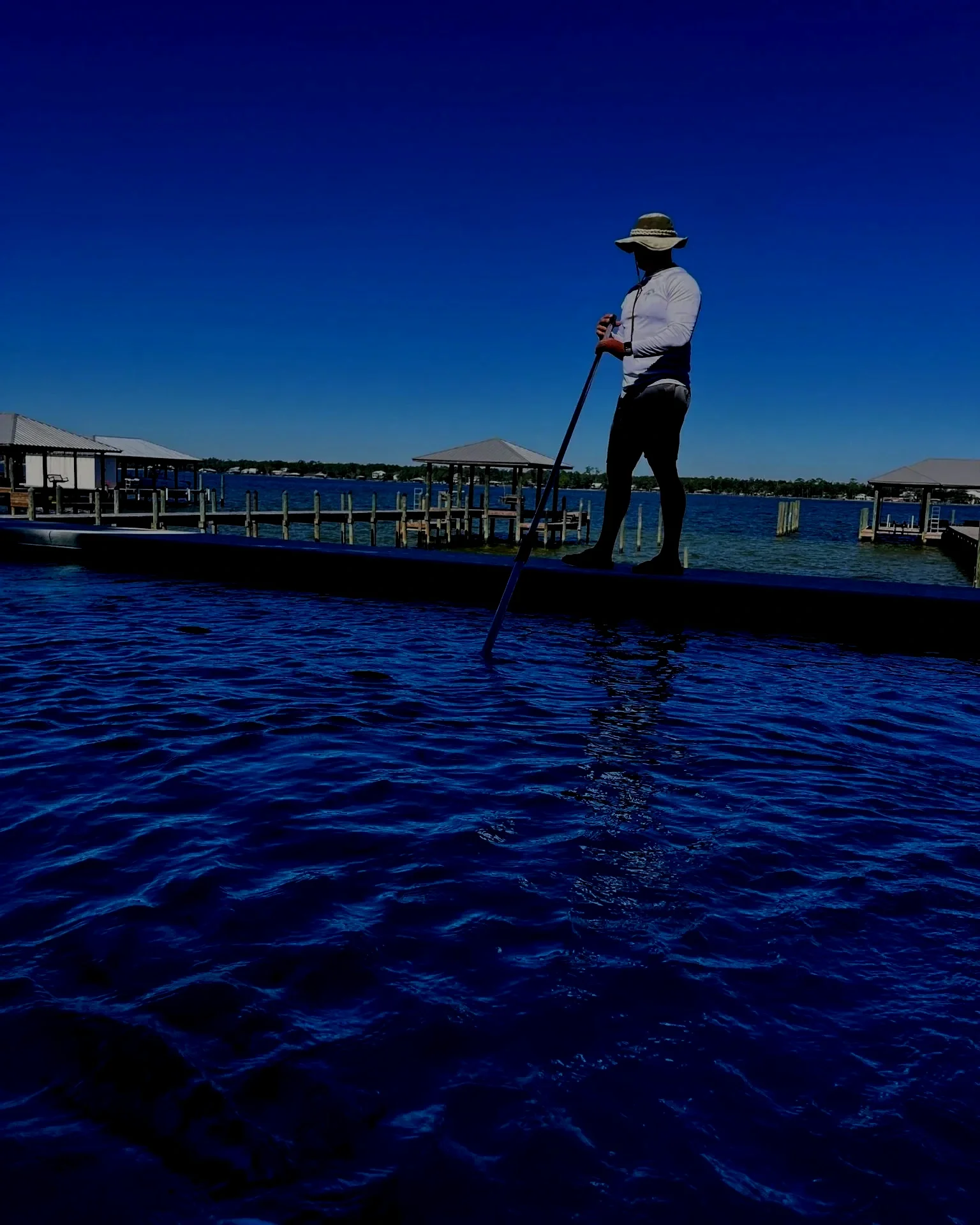 Person wearing a sun hat and gripping a pole while standing on a boat in a marina