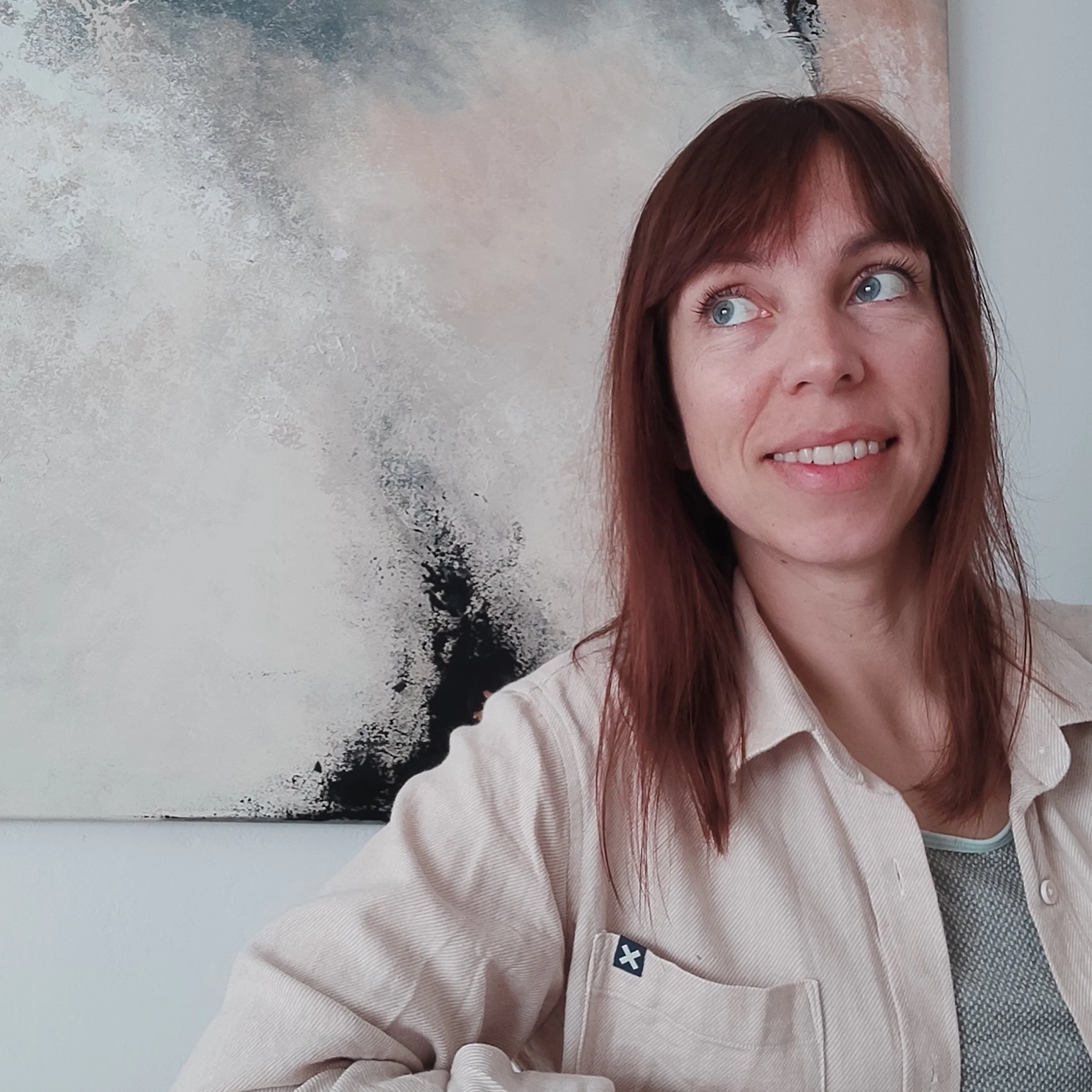 Close-up of a woman with red hair smiling, standing in front of an abstract painting with gray, white, and black textures.