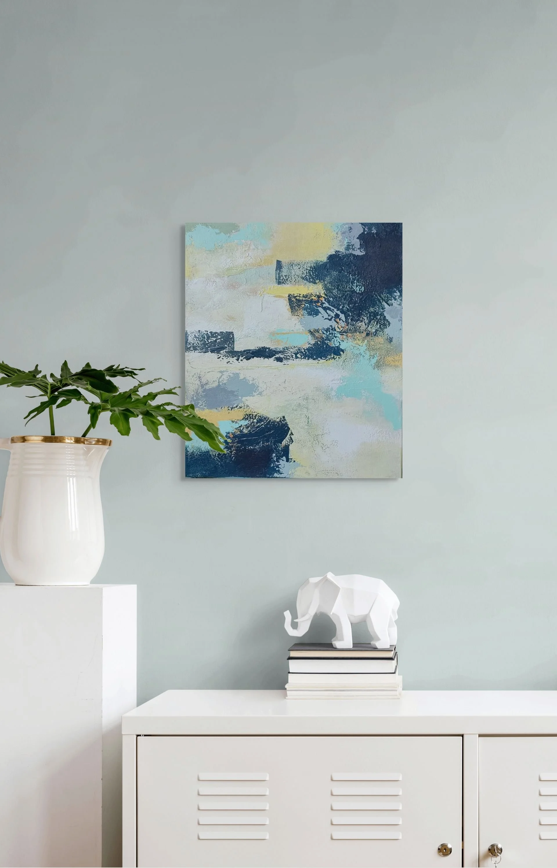 Living room decor with a white cabinet, potted plant, abstract wall art, decorative elephant figurine on books, and a minimalist style.