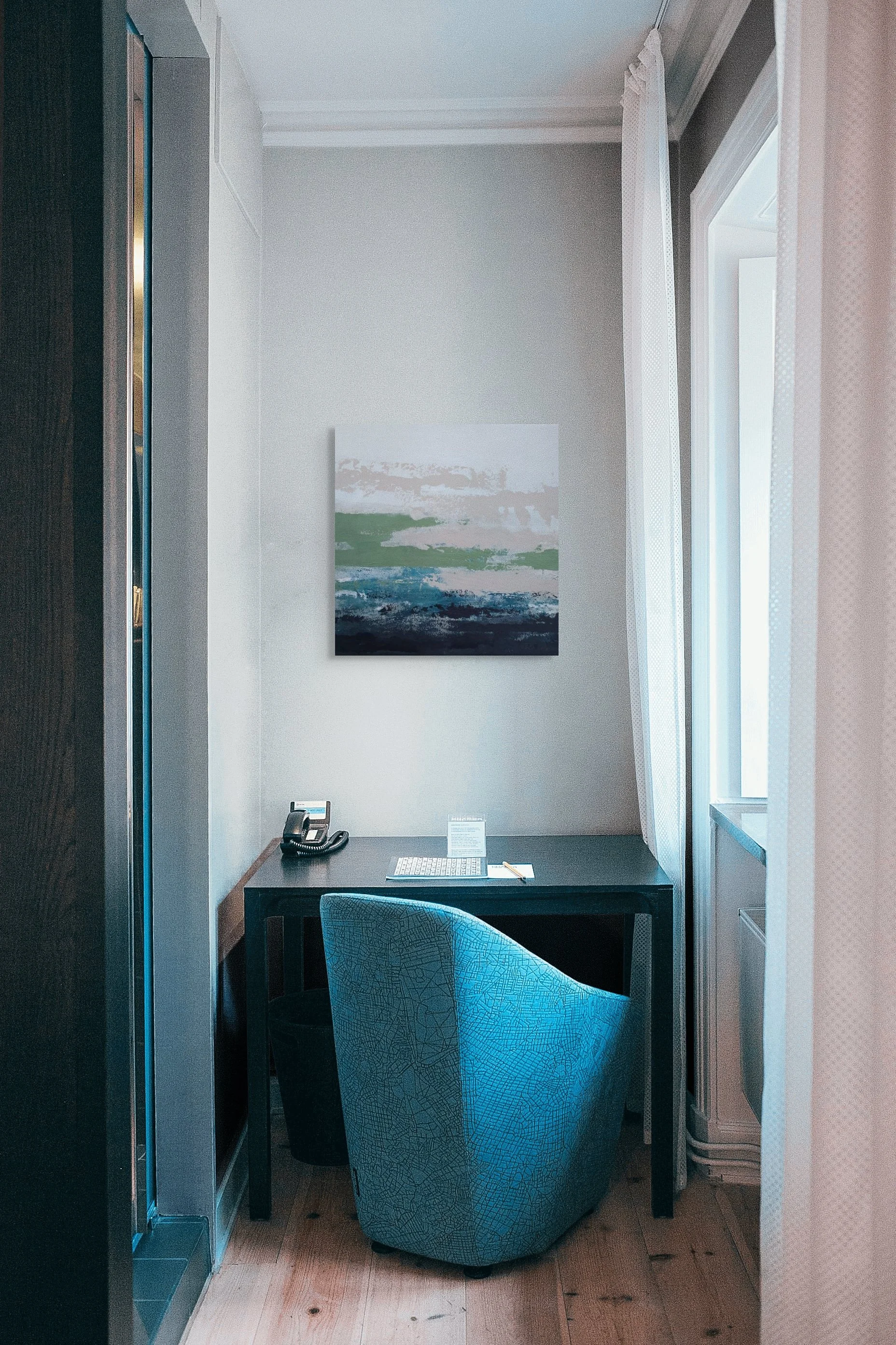 A desk with a chair, a telephone, and papers, positioned in front of a window with curtains, in a room with a painting on the wall.