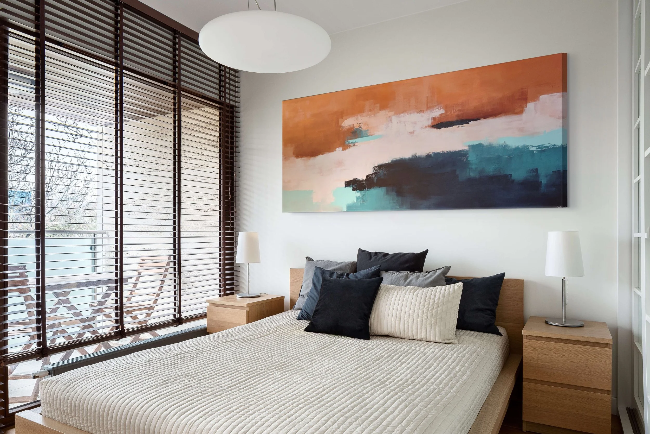 A bedroom with a bed, pillows, and a large abstract painting with blue and terracota tones hanging on the wall. There are two bedside tables with lamps and a window with wooden blinds.