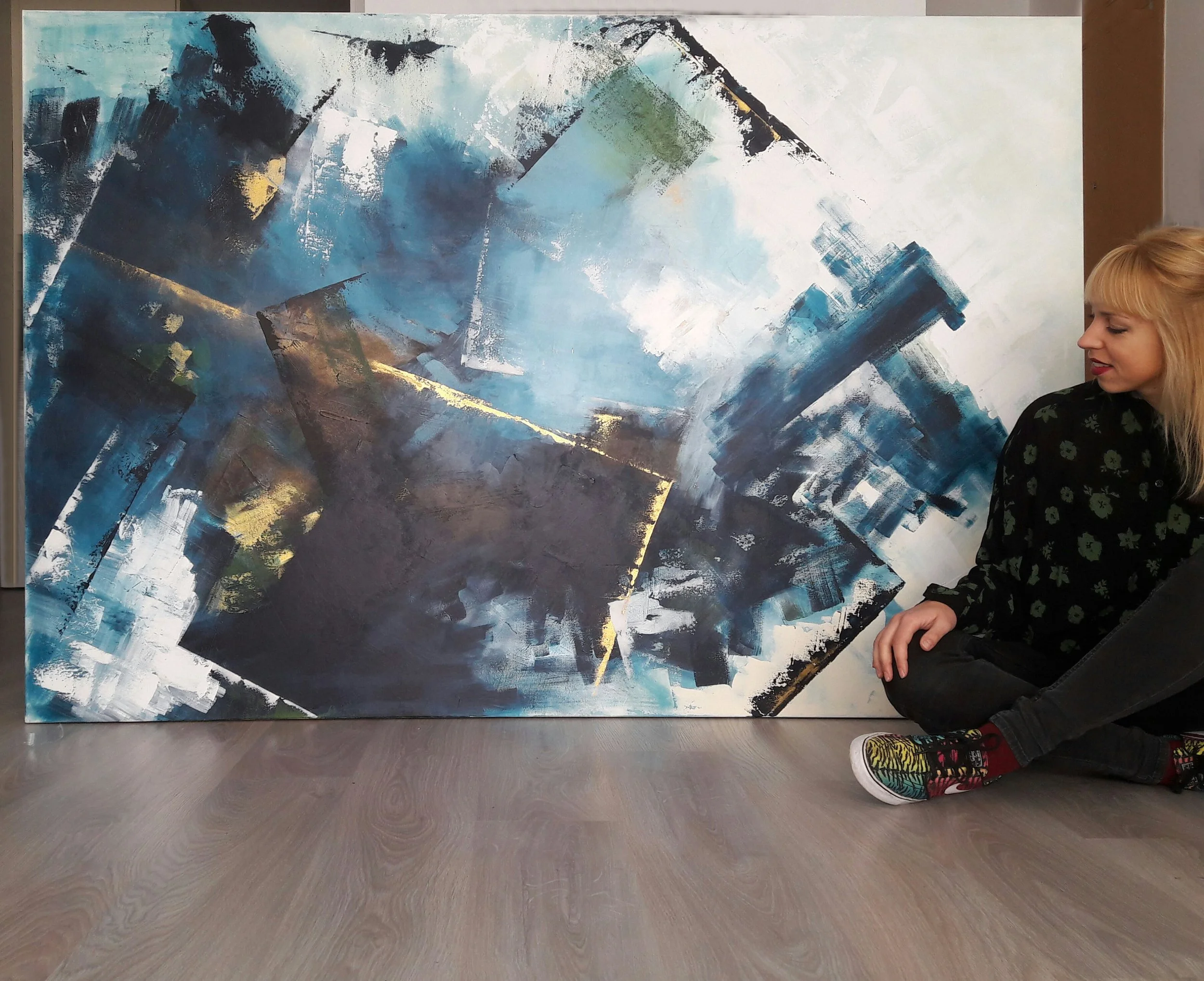 Artista sentada junto a un cuadro abstracto en tonos azules, verdes, negros y dorados, en un espacio interior.