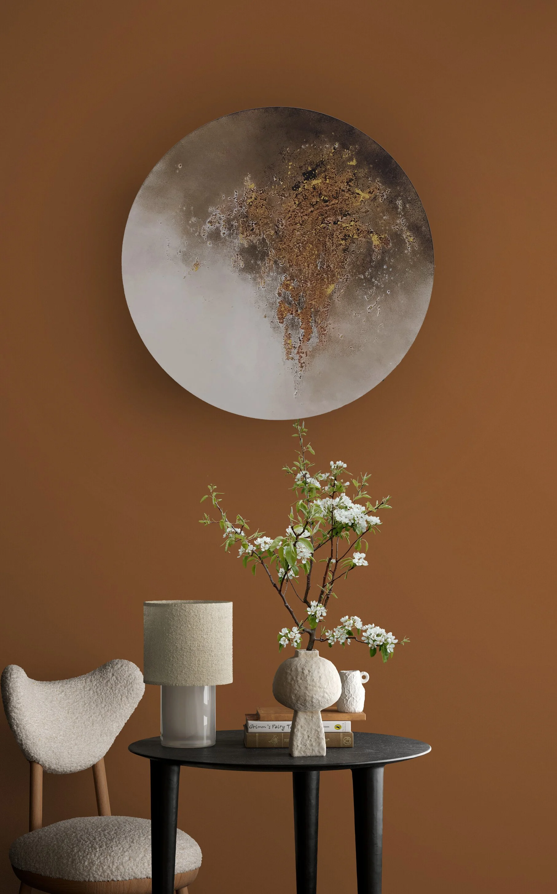 Decorative interior design scene with a black table at the center, holding a white vase with blooming branches, a beige textured lamp, and stacked books, set against a warm brown wall with a large textured circular art piece and minimalist items on t