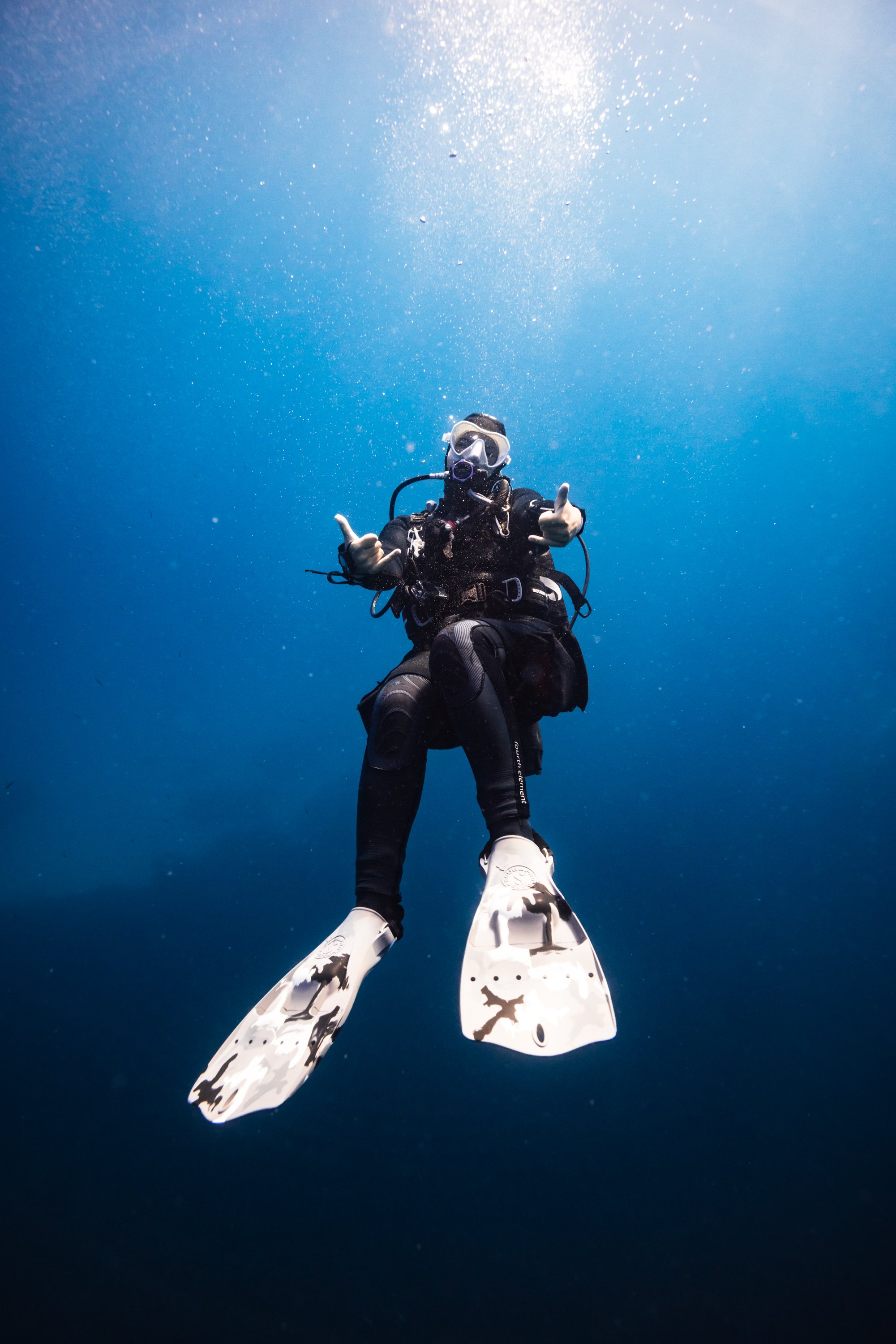 Underwater photograph of a scuba diver wearing a black wetsuit, mask, and oxygen tank, giving a thumbs-up gesture, with a clear blue ocean background and sunlight filtering through the water.