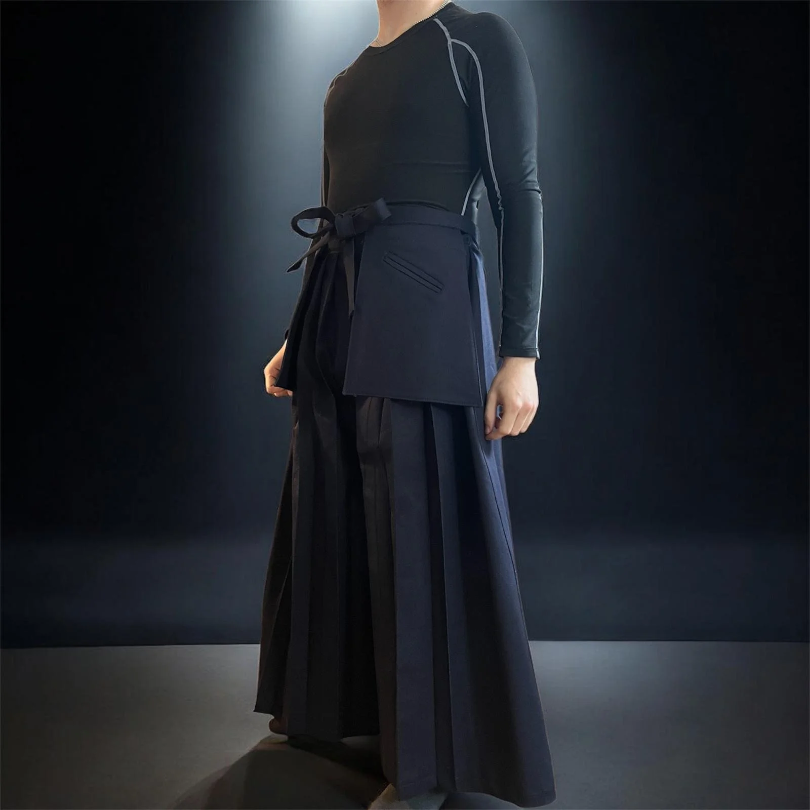 A person dressed in black, wearing a long skirt and a fitted top, standing in a dark environment with a spotlight shining behind them.