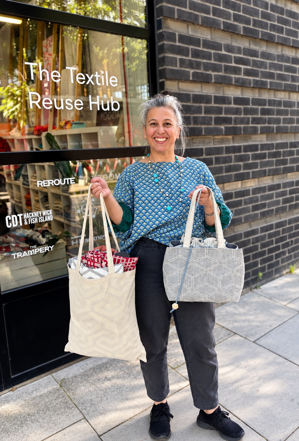 A woman with gray hair smiling and holding two reusable shopping bags outside a store called The Textile Reuse Hub. The woman is wearing a blue patterned top, black pants, and black shoes. The store window behind her displays textiles and textile-related signage.