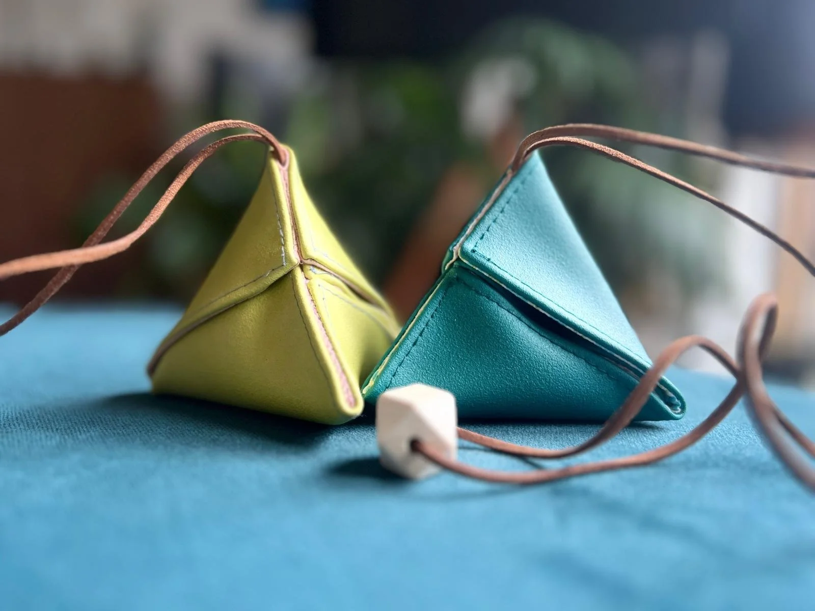 Two colorful pyramid-shaped leather pouches, one yellow and one blue, with brown drawstrings and a small white cube on a blue surface.