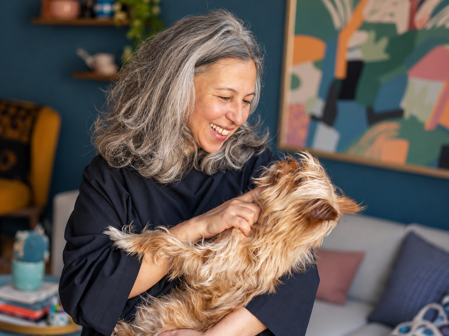 A smiling woman with gray, curly hair holding a small, fluffy, light brown dog, inside a colorful living room.