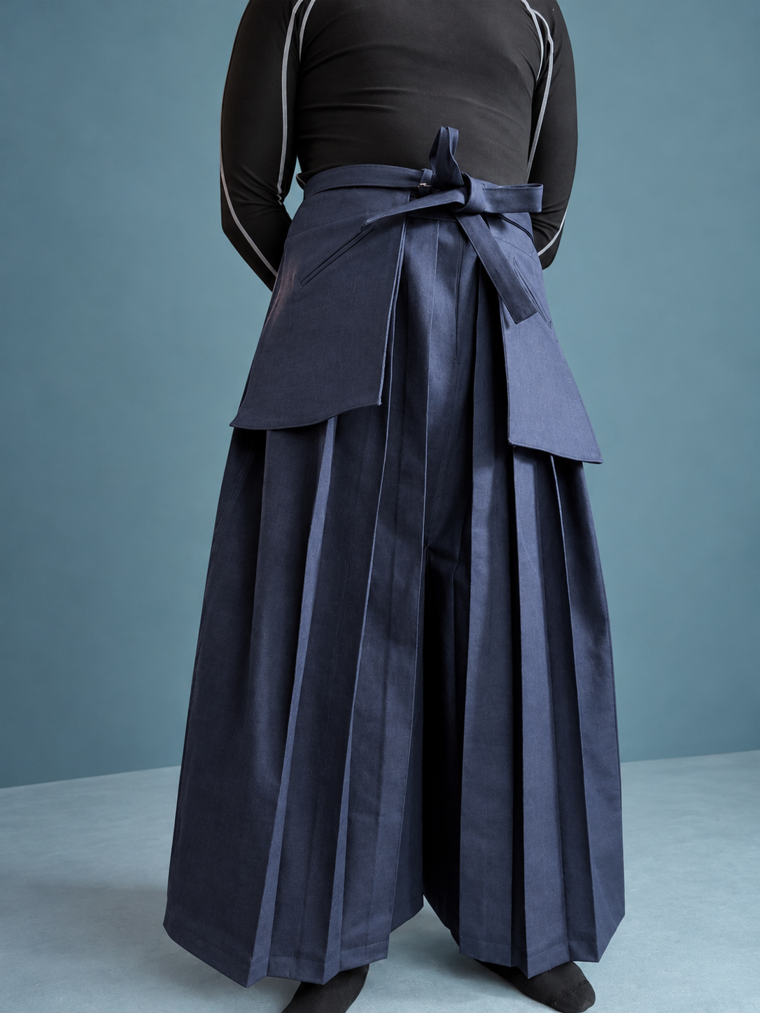 A person wearing a black long-sleeve top and large dark blue pleated pants with a matching apron tied around the waist.