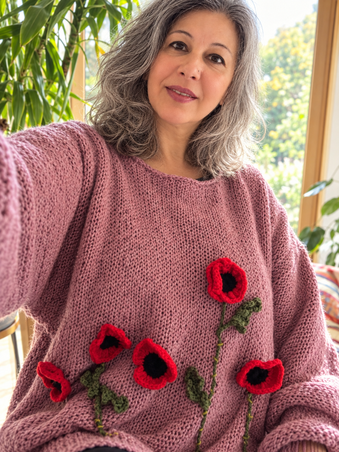 A woman with gray hair taking a selfie inside a sunlit room with large windows and plants, wearing a pink knitted sweater decorated with red poppy flowers.