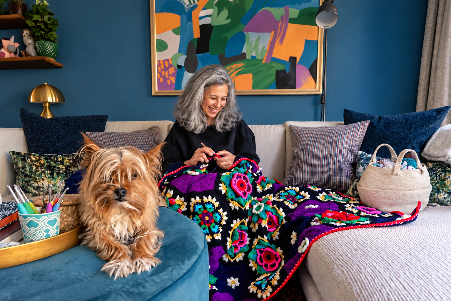 A woman with gray hair sitting on a sofa smiling and knitting, with a large colorful quilt on her lap, next to a fluffy dog lying on a round blue stool, in a cozy living room with blue walls, artwork, a shelf with toys, and decorative pillows.