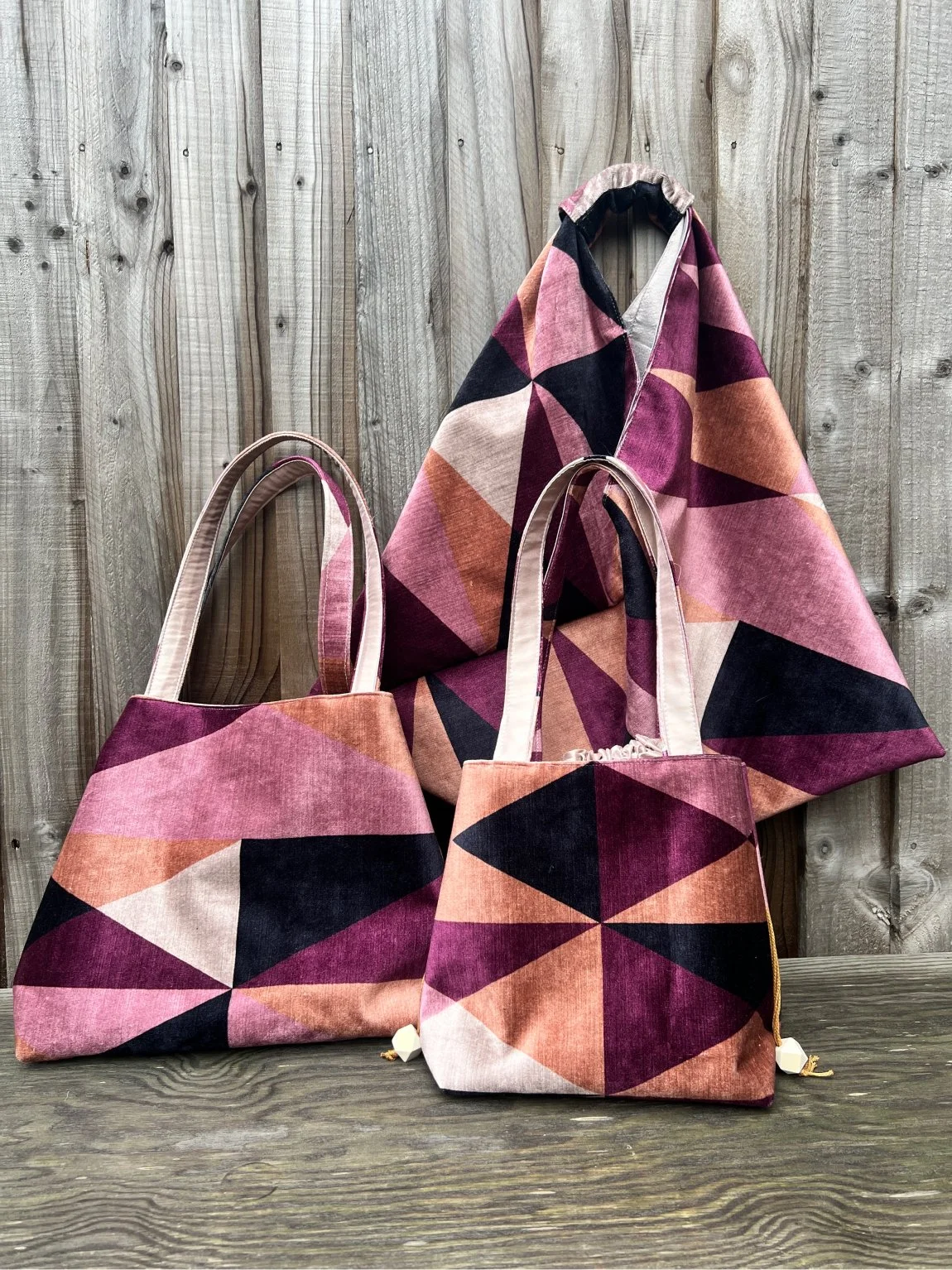 Set of three bags with geometric patterns and pink, beige, black, and peach colors. One bag is hanging on a hook, and two are placed on a wooden surface in front of a wooden wall.
