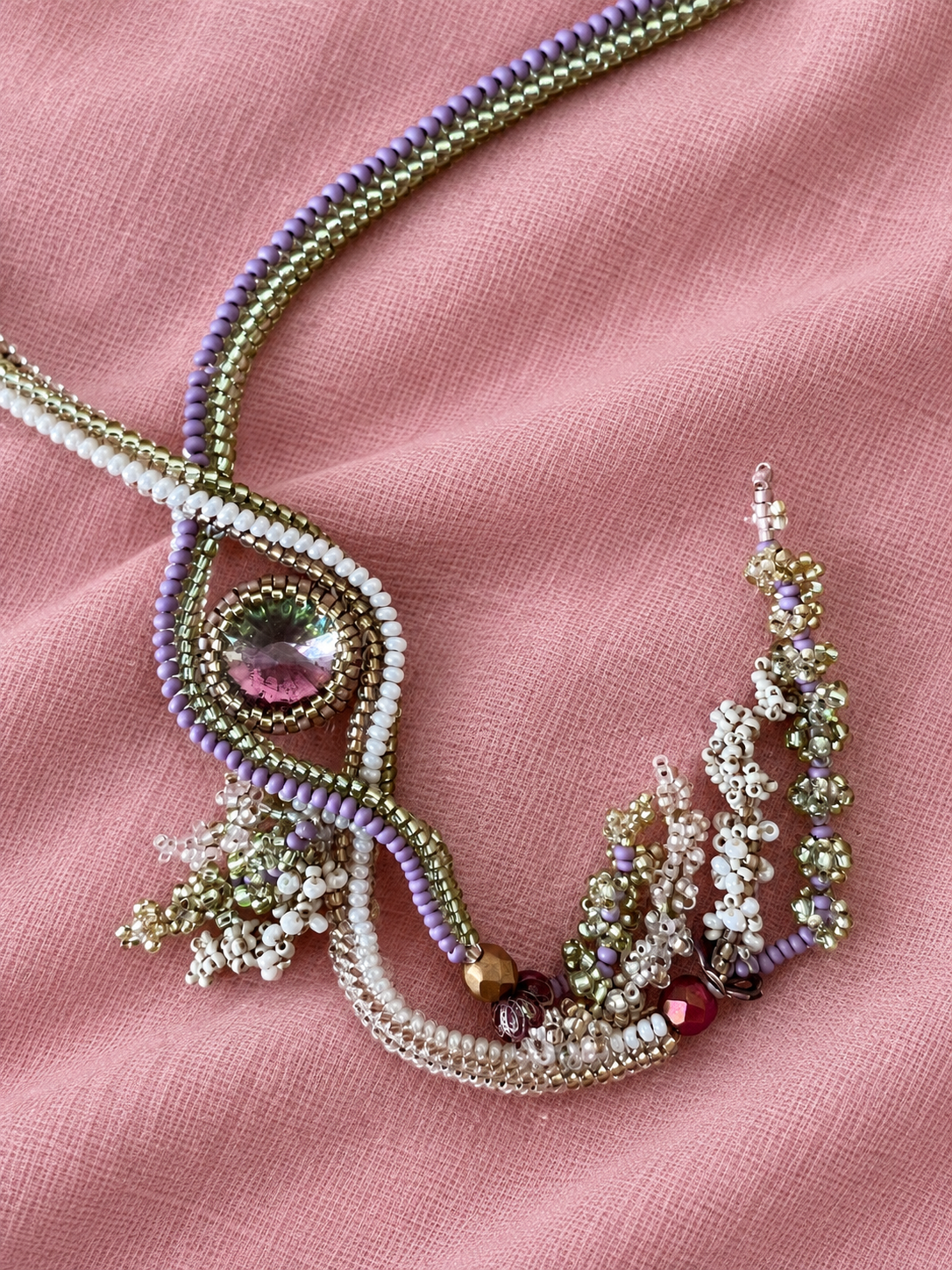 Close-up of a colorful beaded necklace and earrings on pink fabric background. The necklace features purple, white, and gold beads with a central pendant with a pink gemstone. Matching beaded earrings are also shown.