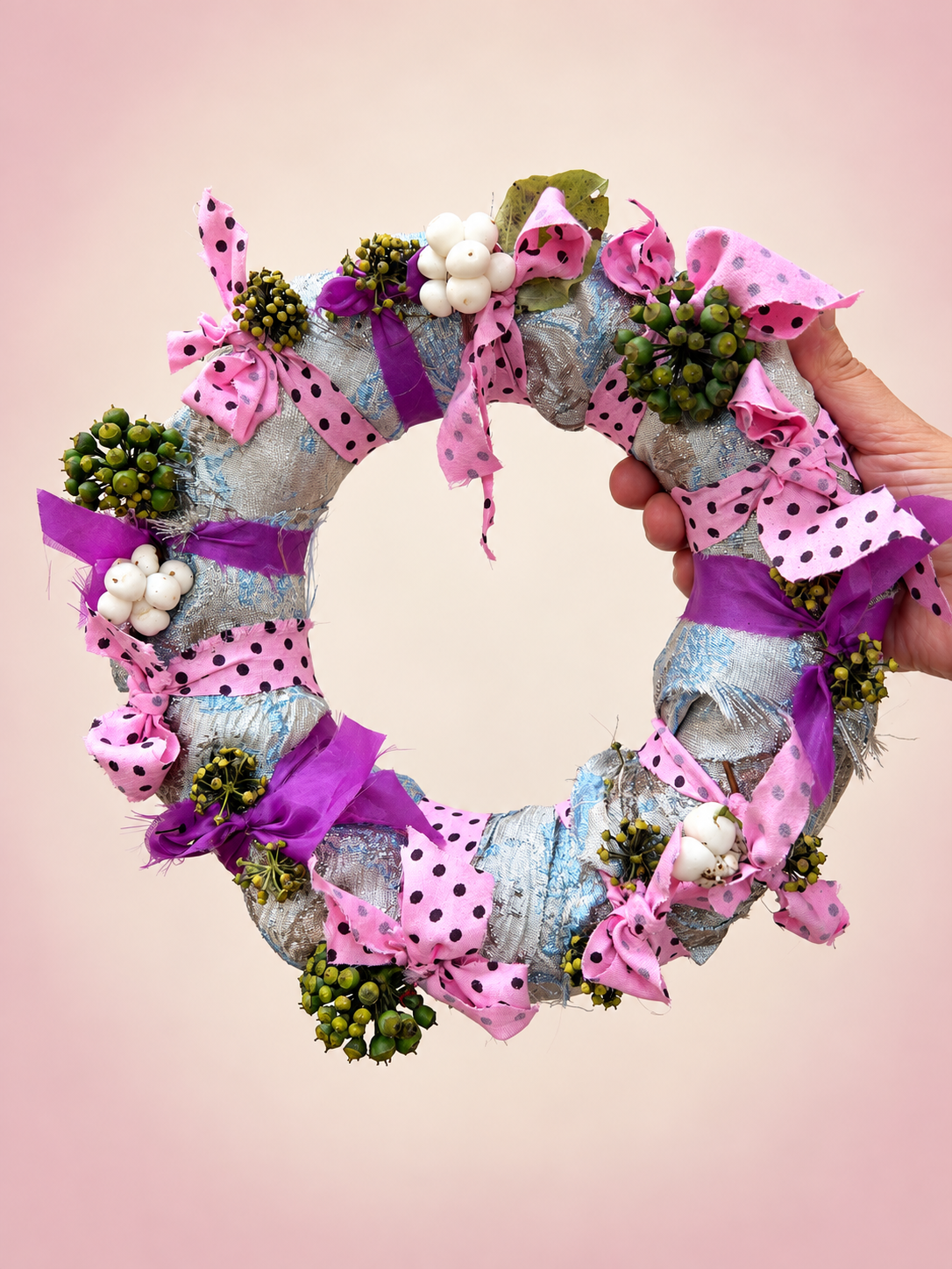 A hand holding a decorative wreath made of fabric, ribbons, pink polka dot bows, small white berries, and greenery.