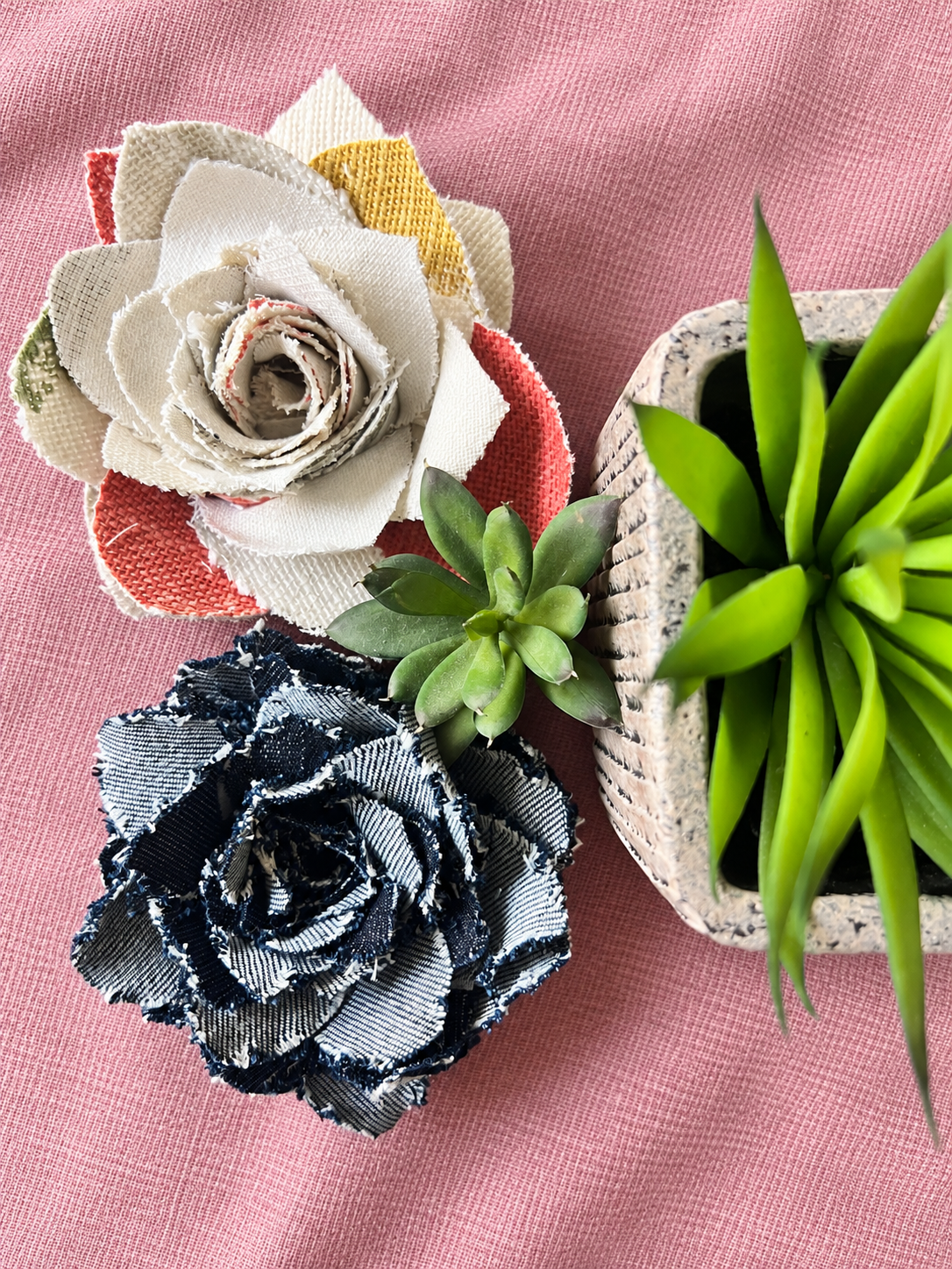 Decorative fabric flowers and potted succulents on a pink textured surface.
