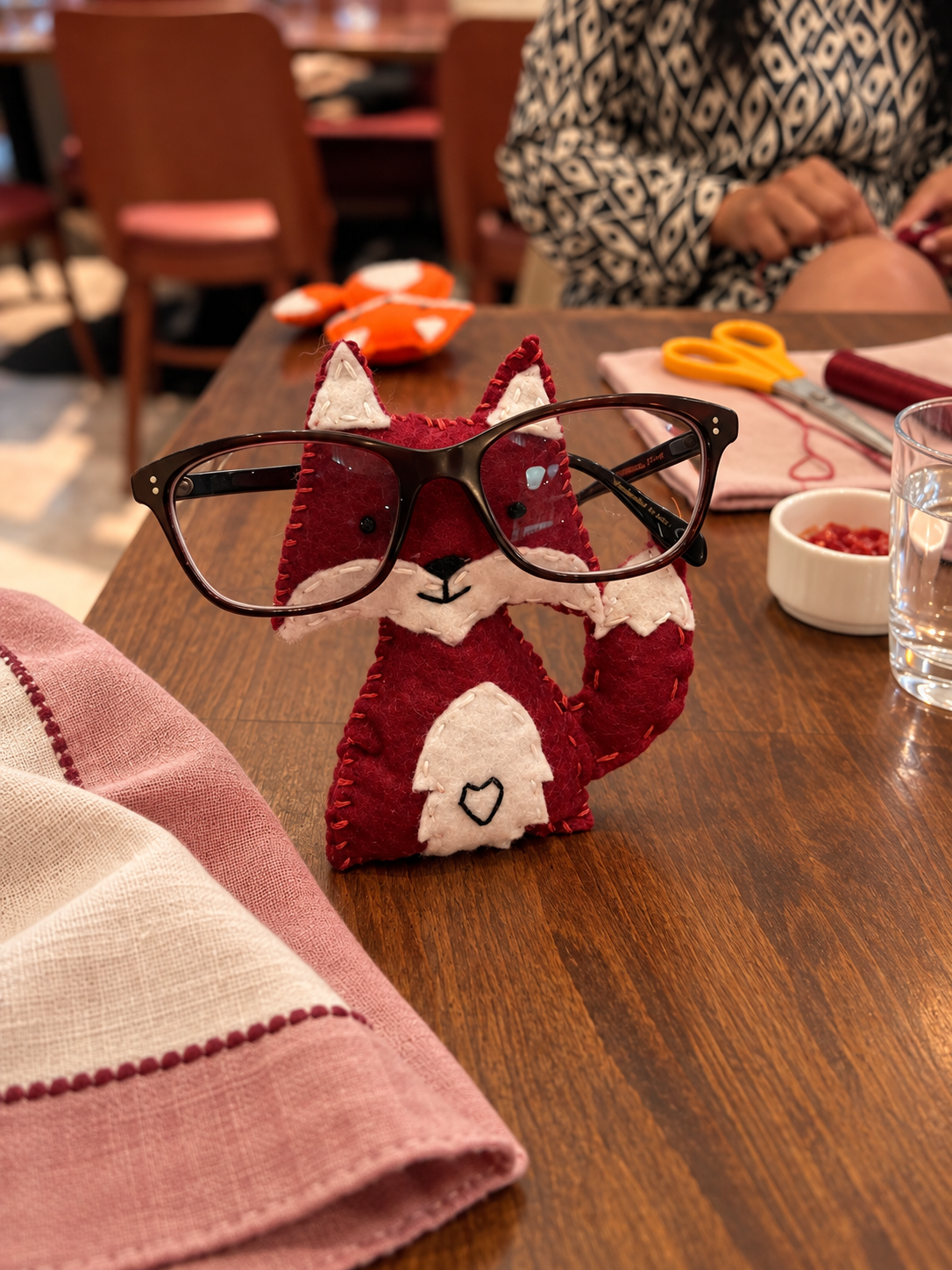A felt plush fox wearing black glasses, sitting on a wooden table in a dining setting. The fox has a red and white design with stitched details.