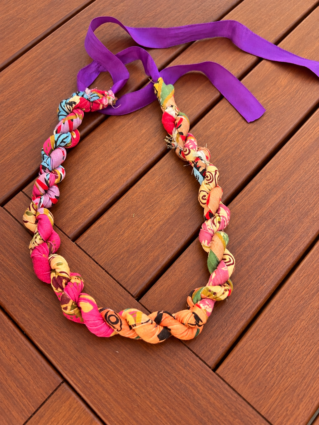 Colorful fabric braided lei on a wooden surface with a purple ribbon.