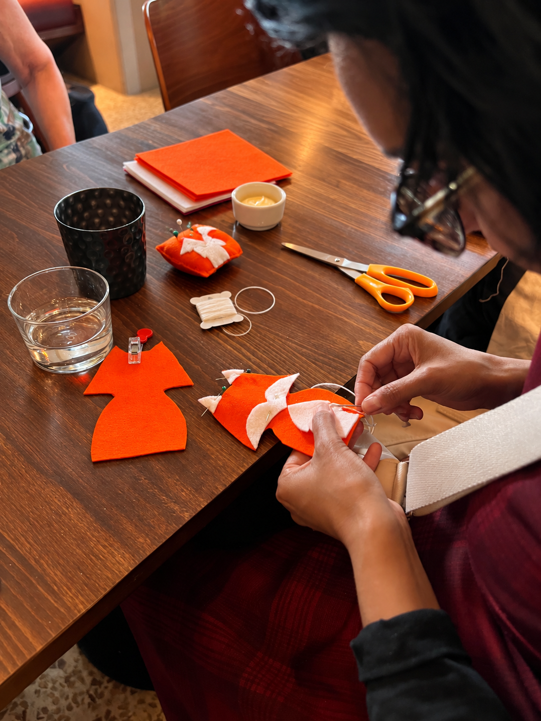 Person sewing small orange felt Christmas stockings with white felt accents at a wooden table, with sewing tools, scissors, and craft supplies scattered around.