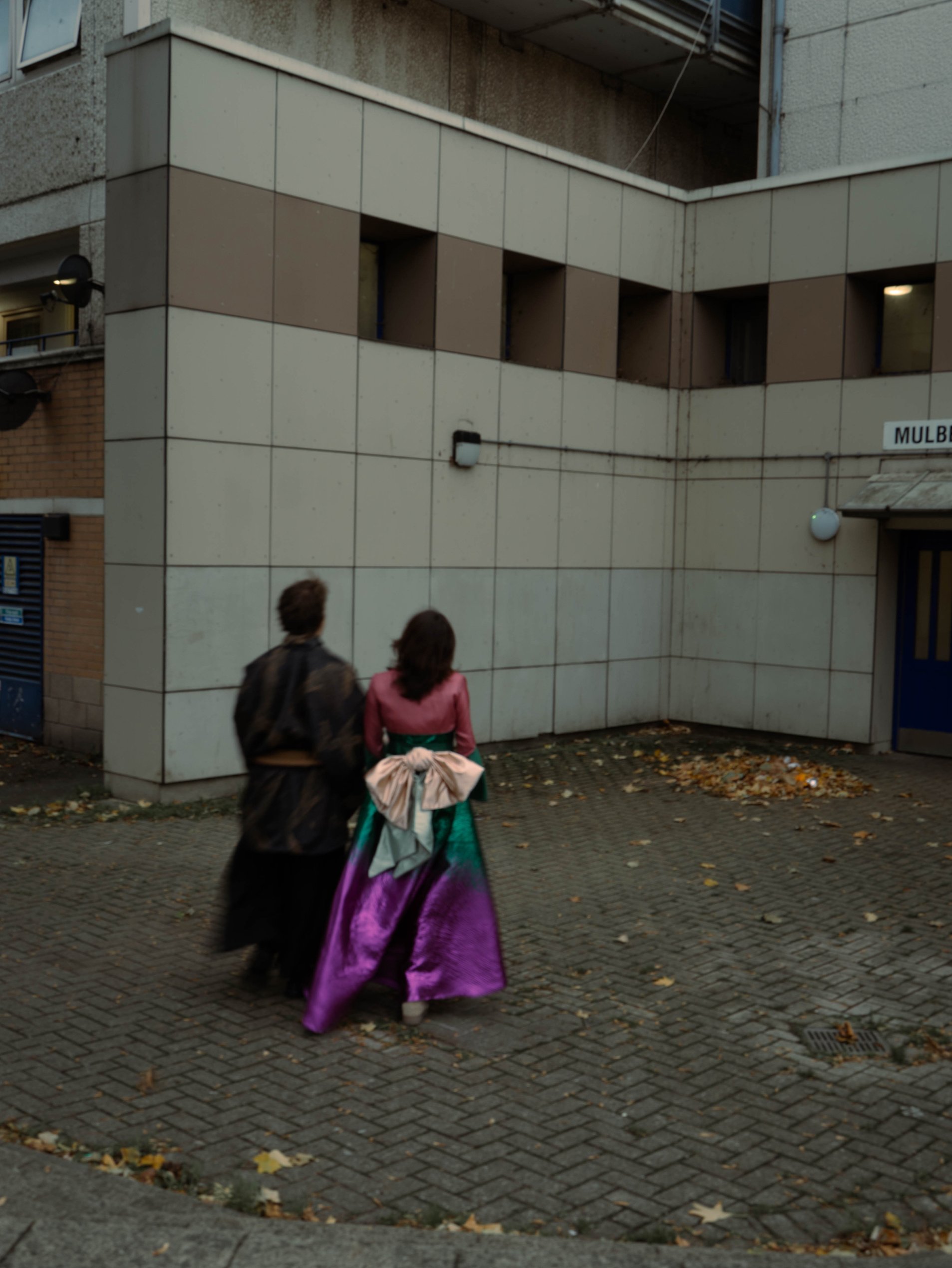 Two people walking on a city sidewalk, one wearing an elaborate purple and green costume, possibly for a costume event or festival, with fall leaves scattered on the ground, and a modern building in the background.