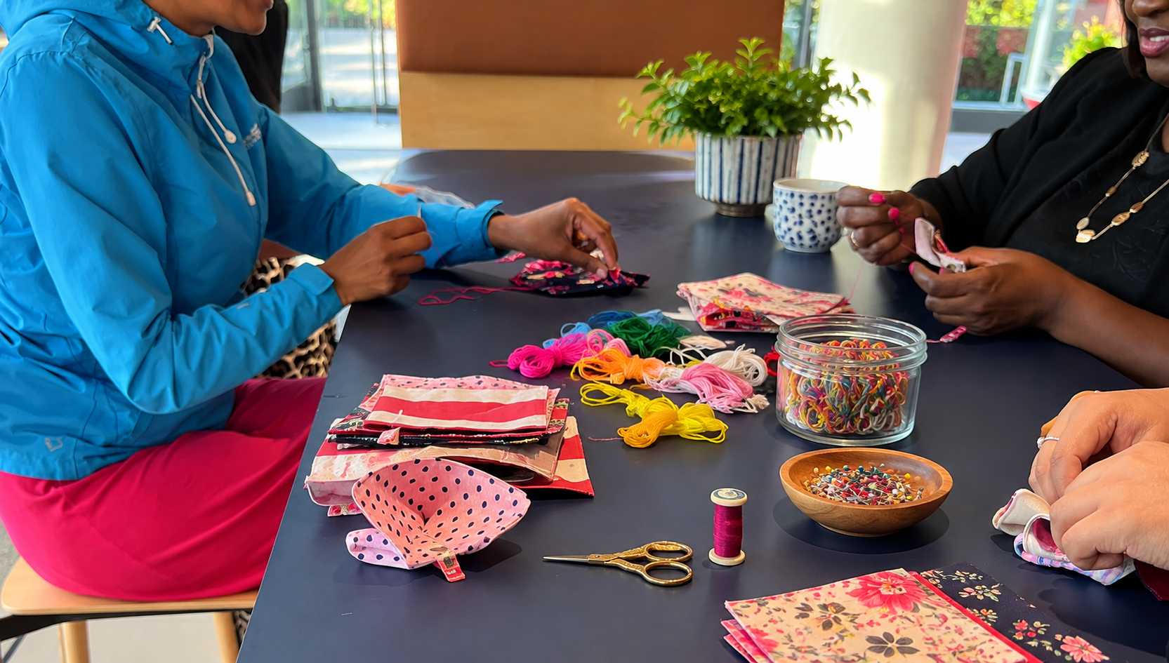Two women sewing with colorful embroidery floss and fabric on a dark table, surrounded by sewing supplies including scissors, pins, fabric pieces, and a container of multi-colored rubber bands.