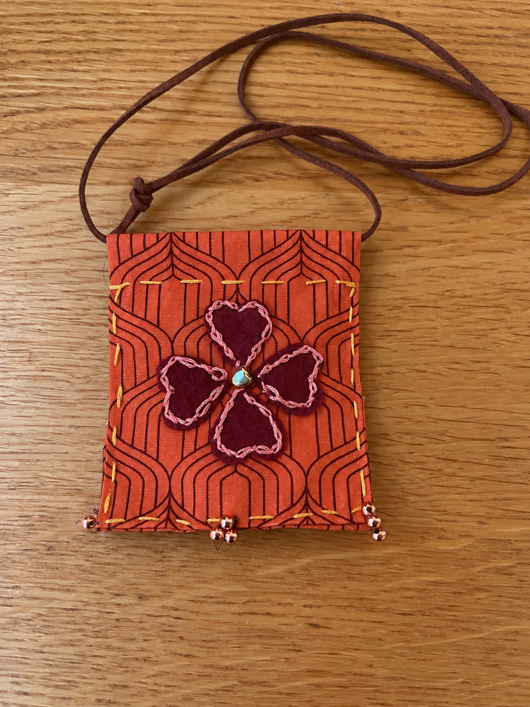 A small fabric bag with a leather strap, decorated with a felt shamrock flower with pink stitched edges and a blue bead at the center, on a wooden surface.