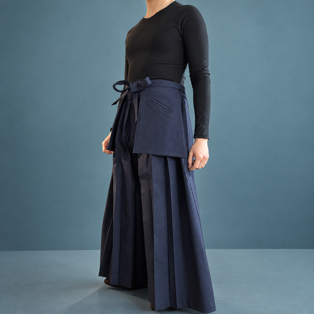 A woman wearing a black long-sleeve shirt and wide-legged navy pants with a matching navy apron tied at the waist, standing against a plain teal background.