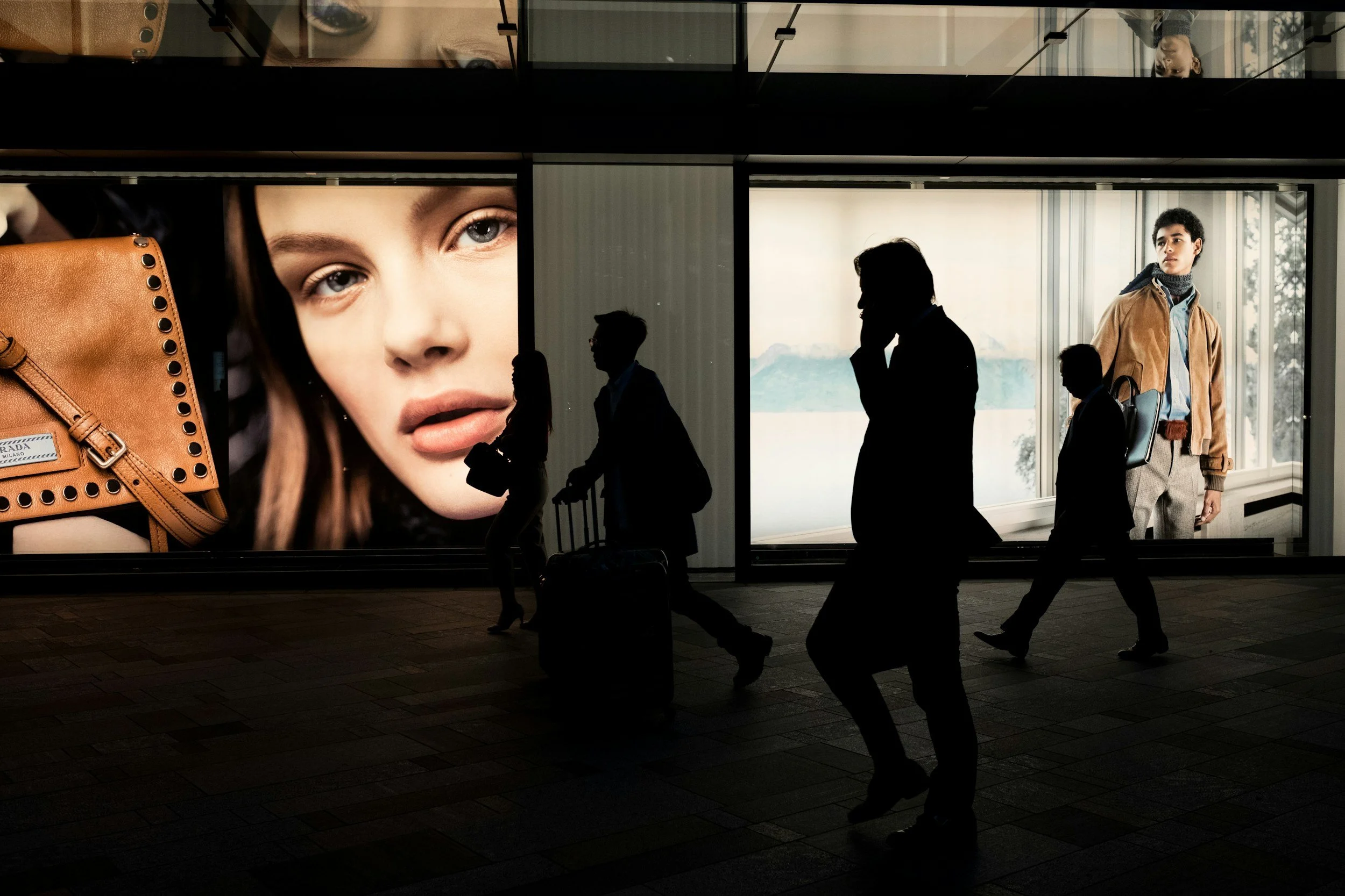Consumer/Retail -  Silhouettes de personnes marchant dans une gare ou un aéroport devant une grande affiche publicitaire montrant un visage d'une femme avec des yeux bleus et un sac à main de couleur camel.