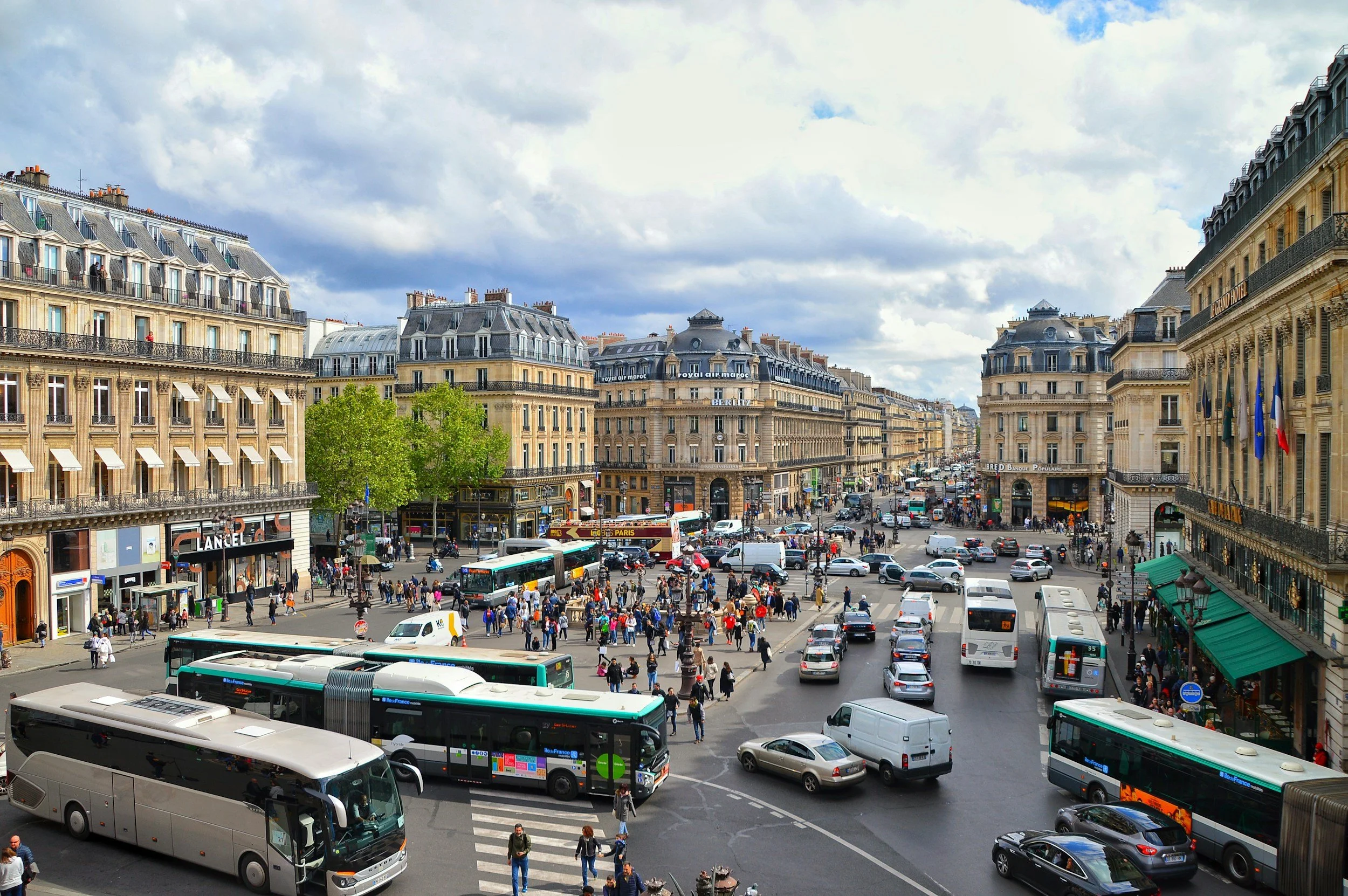 Vue d'une place animée à Paris avec des bâtiments haussmanniens, des bus, des voitures et beaucoup de piétons, par un jour nuageux.