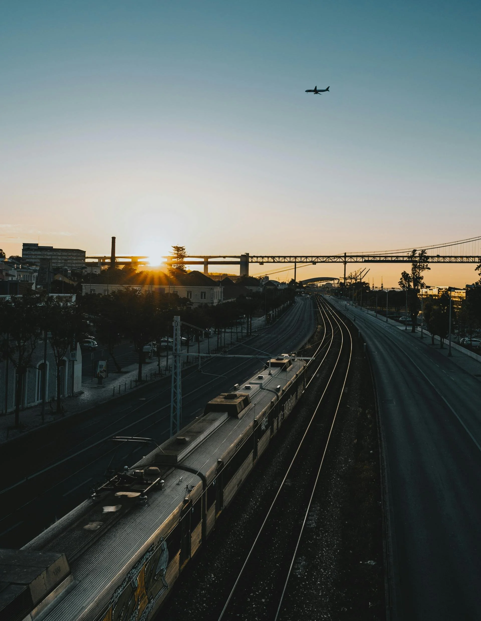 Un train circule sur le rail au coucher du soleil dans une zone urbaine, avec un pont au loin et un avion volant dans le ciel.