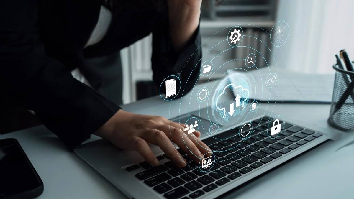 Main tenant un ordinateur portable avec une main en train de toucher le clavier. Des icônes technologiques et de cloud computing sont projetées en hologramme au-dessus du clavier, représentant des données, le stockage en ligne, la sécurité, et la gestion informatique.